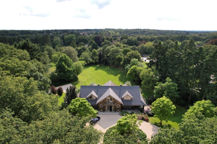 Luchtfoto van een groot, bruin houten Finnhouse met grijs dak, omringd door groene bomen en een verzorgde tuin. Auto op de markt, een bos strekt zich uit aan de horizon. - Finnhouse