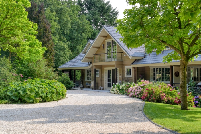 Een betoverend huis met puntdak omgeven door weelderig groen en bloeiende bloemen, een grindpad leidt naar de entree. Grote bomen en struiken omlijsten de tuin voor een rustige en natuurlijke sfeer. - Finnhouse