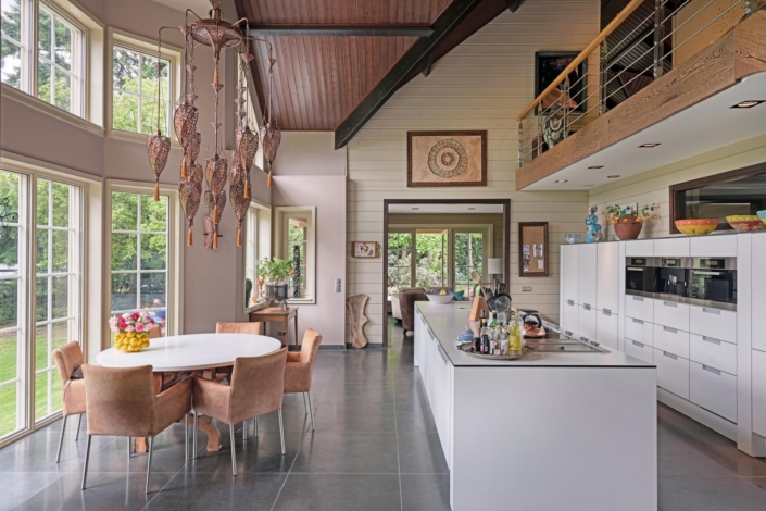 Ruime, moderne keuken met ronde eettafel en bruine stoelen. Grote ramen en een strak wit kookeiland. Hoog plafond met balkon, kunst en een kroonluchter bij de tafel. - Finnhouse