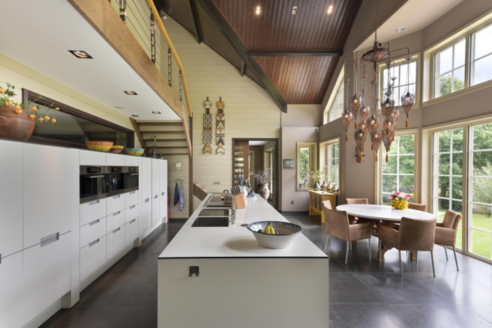 Ruime moderne keuken met hoge plafonds en grote ramen, voorzien van een witte kookeiland, feitelijke apparatuur, eetgedeelte met ronde tafel en stoelen. Veel natuurlijk licht en uitzicht op groen. - Finnhouse