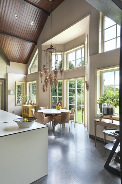 Ruime eetkamer met hoge plafonds en grote ramen die natuurlijk licht binnenlaten. Ronde tafel met beige stoelen, sierlijke kroonluchter, planten als decoratie en uitzicht op groene tuin buiten. - Finnhouse