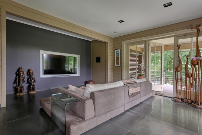 Moderne woonkamer met grote grijze hoekbank tegenover wandgemonteerde tv, glazen deuren naar balkon met buiten groen. Houten sculpturen en decoratie sieren de ruimte met tegelvloer en neutrale muren. - Finnhouse