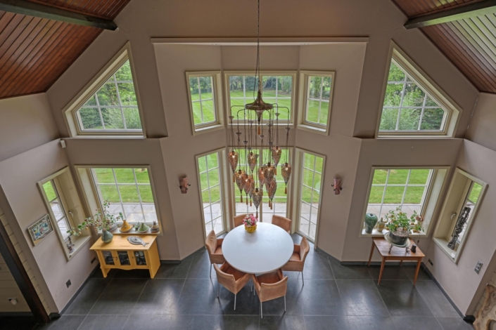 Luchtfoto van een eetkamer met ronde witte tafel en zes stoelen bij Finnhouse.nl. Grote ramen en hoog plafond zorgen voor veel natuurlijk licht, met stijlvolle kroonluchter en decoratieve elementen. - Finnhouse