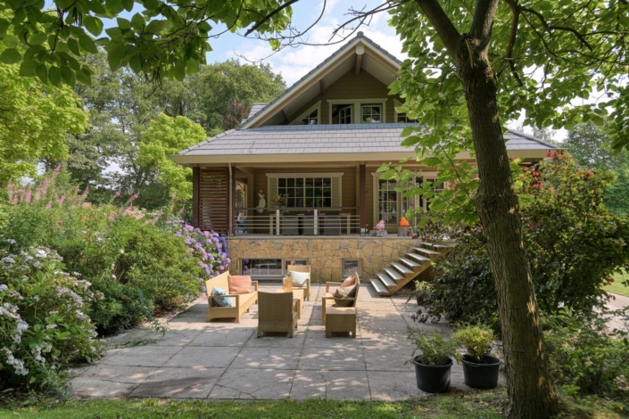 Charmante Finnhouse woning met stenen en houten gevel, omringd door weelderig groen. Voorporch met trap leidt naar knusse patio met houten meubels en kleurrijke kussens, bewegende door bloeiende struiken en bomen. - Finnhouse