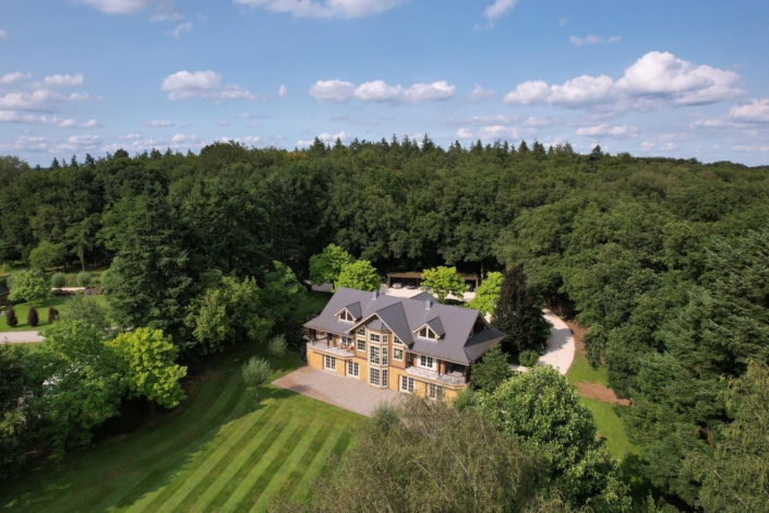 Luchtfoto van een groot binnenkant huis met donker dak en grote ramen, afdaling door weelderig groen en bomen. Goed onderhouden gazon met duidelijke strepen en oprit door het bos. Deels bewolkte hemel. - Finnhouse