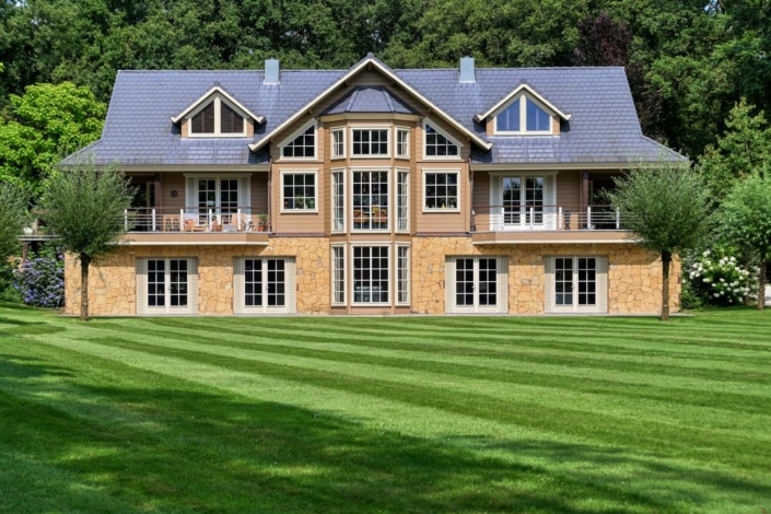 Modern houten huis van Finnhouse met grote raampartijen en duurzaam ontwerp, buitenkant door een groene, goed onderhouden tuin met bomen, onder een strakke leistengrijze dakbedekking. - Finnhouse