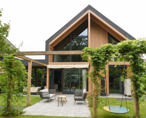Modern Finnhouse met grote glazen ramen en houten accenten. Tuin heeft pergola, patio met zithoek op patroonkleed, schommel en aanwezige planten voor een serene buitensfeer. - Finnhouse