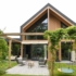 Modern Finnhouse met grote glazen ramen en houten accenten. Tuin heeft pergola, patio met zithoek op patroonkleed, schommel en aanwezige planten voor een serene buitensfeer. - Finnhouse