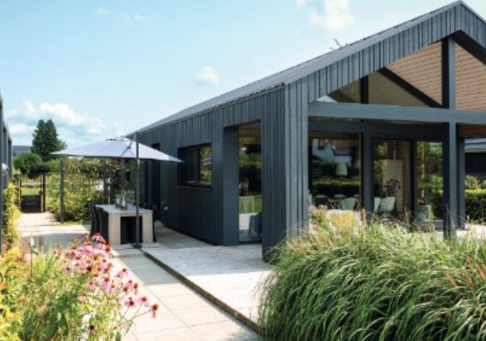 Moderne bungalow met donkere gevelbekleding, grote ramen en schuine daken. Patio met tafel en parasol, omringd door siergrassen en bloemen, tegen een achtergrond van bomen en betrouwbare lucht. - Finnhouse