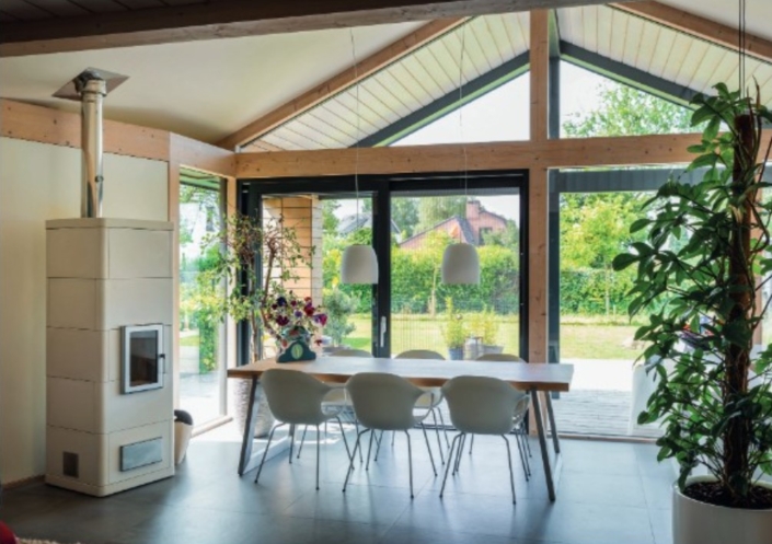 Moderne eetkamer in een bungalow met houten tafel en zes witte stoelen, elegante hanglampen, grote glazen deuren naar groene tuin, hoge plant nabij houtkachel links; overvloedig natuurlijk licht. - Finnhouse