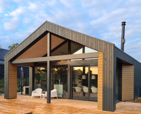 Moderne bungalow met houten exterieur en grote glazen ramen, gelegen in een groene vloeibare omgeving. Twee witte stoelen op houten terras, onder deels bewolkte lucht en onderaan een parasol. - Finnhouse