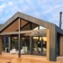 Moderne bungalow met houten exterieur en grote glazen ramen, gelegen in een groene vloeibare omgeving. Twee witte stoelen op houten terras, onder deels bewolkte lucht en onderaan een parasol. - Finnhouse