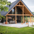 Moderne schuurwoning van Finnhouse met donker, steil dak en houten balken, omringd door ruim houten terras met tuinmeubilair en planten. Uitzicht op groen grasveld en open landschap onder bewolkte hemel. - Finnhouse