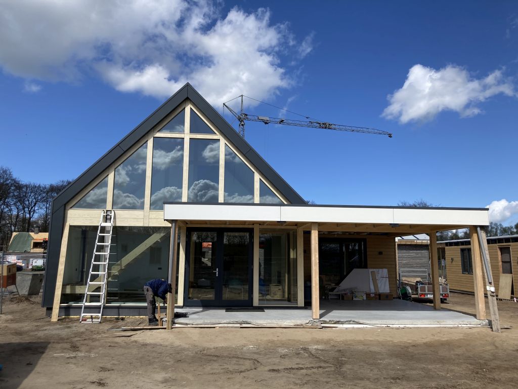 Een moderne Finnhouse in aanbouw met grote glazen panelen en een stalen dak; een arbeider op de ladder, terwijl een kraan hoog oprijst tegen de deels bewolkte hemel. - Finnhouse