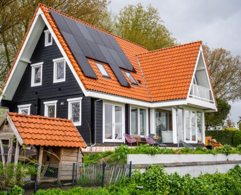 Een moderne tweeverdiepingswoning in Logbouw 4158 stijl, met zwartwitte gevelbekleding en een steil rood pannendak met zonnepanelen; beneden door weelderig groen en houten schuurtje. - Finnhouse