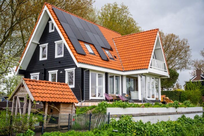 Een moderne tweeverdiepingswoning in Logbouw 4158 stijl, met zwartwitte gevelbekleding en een steil rood pannendak met zonnepanelen; beneden door weelderig groen en houten schuurtje. - Finnhouse