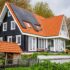 Een moderne tweeverdiepingswoning in Logbouw 4158 stijl, met zwartwitte gevelbekleding en een steil rood pannendak met zonnepanelen; beneden door weelderig groen en houten schuurtje. - Finnhouse