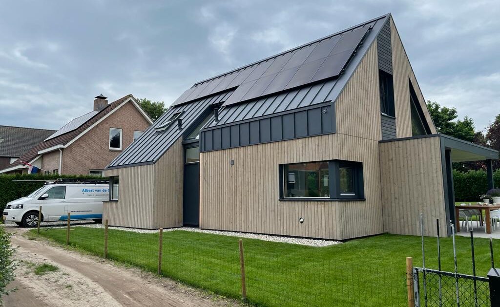 Moderne tweeverdiepingen Finnhouse met hellend dak, bekleed met licht hout en voorzien van zonnepanelen. Omgeven door verzorgde tuin; witte bestelwagen staat geparkeerd onder bewolkte lucht. - Finnhouse