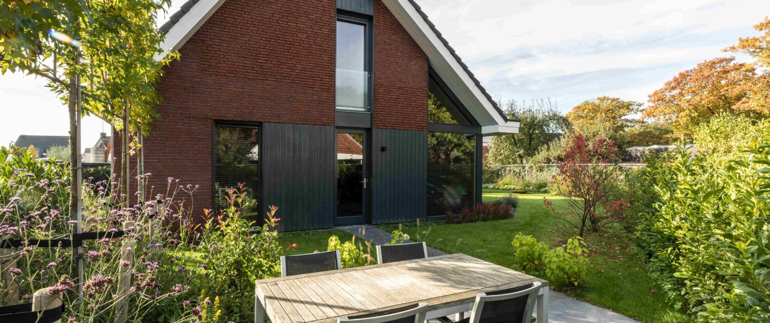 Een modern Finnhouse met steil dak, grote ramen en verticale donkere panelen. Op het terras staan een houten eettafel met stoelen, omringd door groene planten en bomen. - Finnhouse