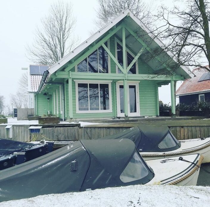 Een groene houten Finnhouse met grote ramen staat bij een besneeuwd dok, naast door kale bomen. Overdekte boten liggen aangemeerd terwijl de winterse lucht boven Oostrum bewolkt is. - Finnhouse