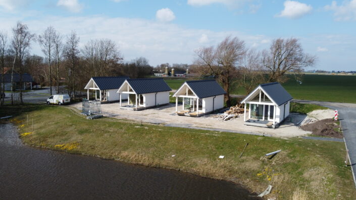 Vier moderne, witte huisjes met donkere daken en veranda's aan een rivier, lijken op een horeca-paviljoen in Oostrum. Nabij zijn bouwmaterialen zichtbaar, met links een busje tussen kale bomen. - Finnhouse