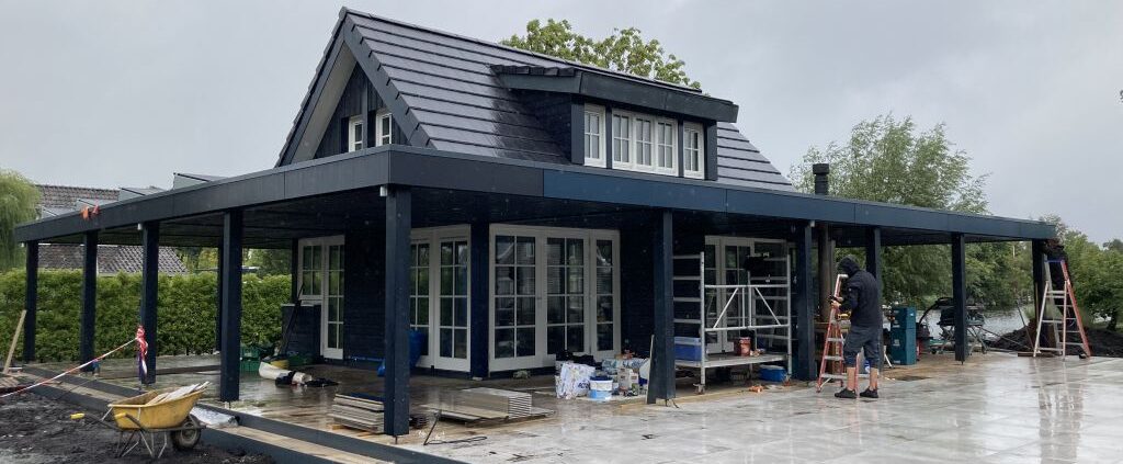 Een houten Finnhouse met donkere gevelbekleding en pannendak, omringd door renovatiematerialen en werklui, op een natte patio. Bewolkte lucht hint naar dynamische omstandigheden tijdens de verbouwing. - Finnhouse