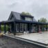 Een houten Finnhouse met donkere gevelbekleding en pannendak, omringd door renovatiematerialen en werklui, op een natte patio. Bewolkte lucht hint naar dynamische omstandigheden tijdens de verbouwing. - Finnhouse