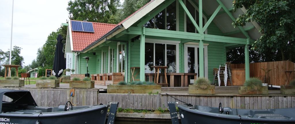 Groene houten Finnhouse met dak en zonnepanelen aan het water, te Oostrum. Kleine donkergrijze bootjes liggen aangemeerd tussen de bomen onder een bewolkte hemel. - Finnhouse