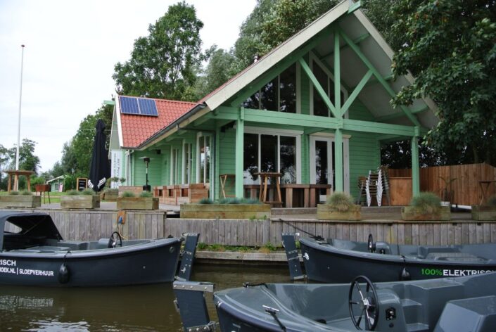 Groene houten Finnhouse met dak en zonnepanelen aan het water, te Oostrum. Kleine donkergrijze bootjes liggen aangemeerd tussen de bomen onder een bewolkte hemel. - Finnhouse
