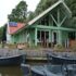 Groene houten Finnhouse met dak en zonnepanelen aan het water, te Oostrum. Kleine donkergrijze bootjes liggen aangemeerd tussen de bomen onder een bewolkte hemel. - Finnhouse