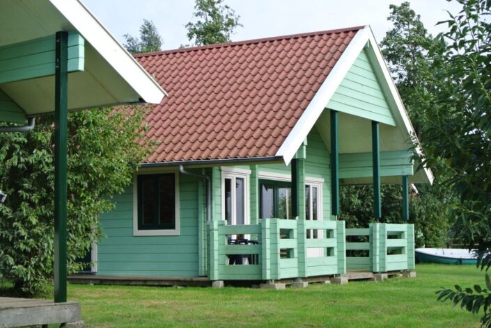 Een charmant, lichtgroen houten huis met rode dakpannen en veranda, achter door gras en bomen in Oostrum. Witte deur en ramen geven dit Horeca paviljoen een gezellige uitstraling. - Finnhouse