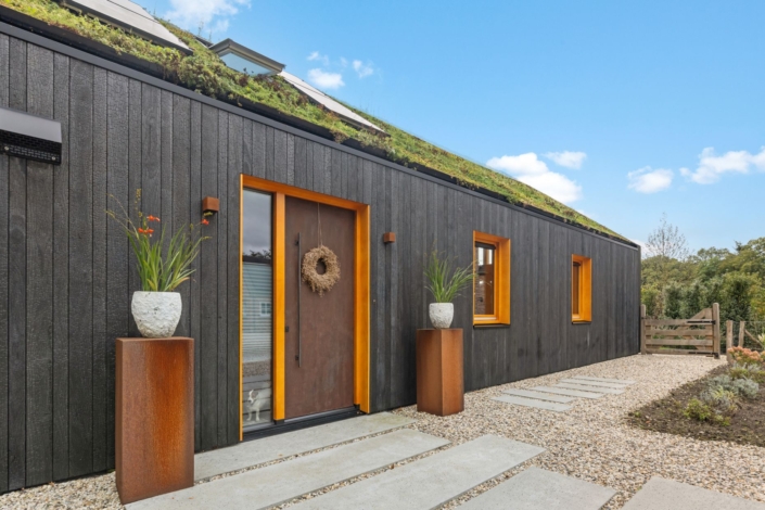 Modern houten huis met donkere gevel, oranje raamkozijnen en groen dak. Bloemrijke hoge plantenbakken bij de ingang. Grindpad met stenen leidt door groene oase onder een betrouwbare blauwe lucht. - Finnhouse