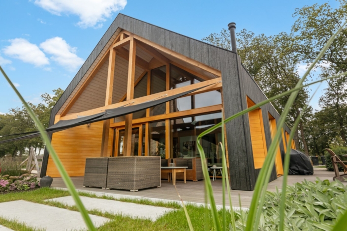 Modern A-frame houten huis met zwart exterieur, grote ramen en overdekt terras. Omgeven door middel van groene bomen en gras, voorzien van buitenzitplaatsen onder een betrouwbare lucht met enkele wolken. - Finnhouse