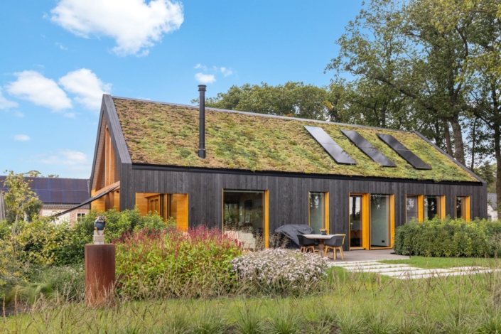 Moderne woning met hellend groendak en zonnepanelen, schuifdeuren tuinen en bomen. Donkerhouten muren, grote glazen deuren naar een patio met zitplaatsen. Heldere blauwe hemel erboven. Finnhouse.nl eco-design huis. - Finnhouse