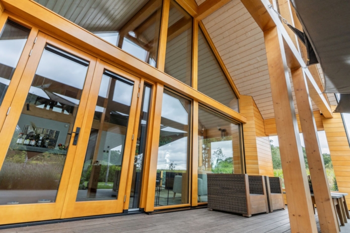 Moderne houten woning met grote glazen deuren en ramen, hoog schuin dak en terras met tuinmeubelen. Omringd door verticale bomen en groen, straalt het een natuurlijke charme uit. Finnhouse.nl. - Finnhouse