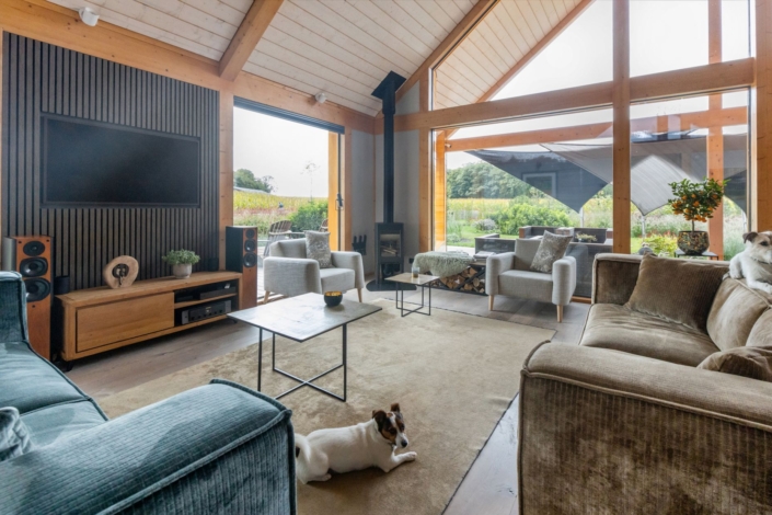 Gezellige woonkamer met grote ramen die uitzicht op groene tuin bieden, bevat twee banken, twee fauteuils, een wandtelevisie en houten balken. Een hond ligt op de vloer naast een salontafel. - Finnhouse