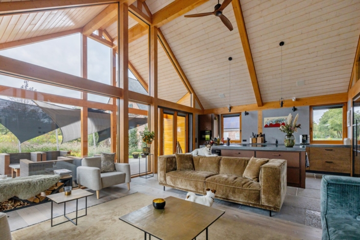 Ruime woonkamer met hoge houten plafonds en grote ramen, ingericht met een comfortabele bank, fauteuils, salontafels en keuken met barkrukken. Een hond ligt op de vloer; groene tuinen zichtbaar buiten. - Finnhouse