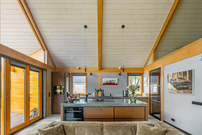 Moderne keuken met gewelfd plafond en houten balken, groot eiland met grijs aanrechtblad, beige bank vooraan, brede ramen en kunstwerken aan de muren. Minimalistische inrichting met natuurlijke verlichting en groenaccenten bieden een luchtige sfeer. - Finnhouse