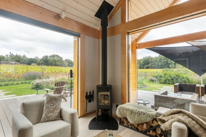 Moderne ruime woonkamer met grote glazen ramen, houtkachel en lichte houten accenten. Uitzicht op groene tuin met vaste vegetatie. Comfortabele fauteuil en buitenzitje versterken de serene sfeer. Finnhouse-ontwerp. - Finnhouse