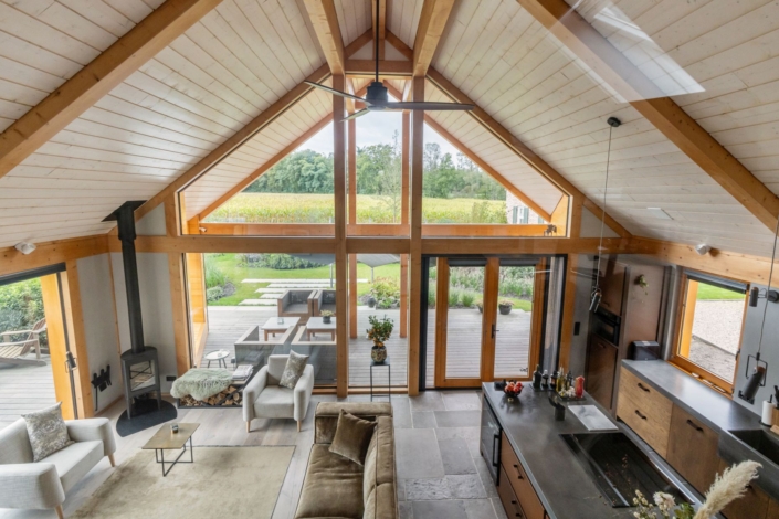 Ruime, moderne woonkamer met hoge houten plafonds en grote ramen die uitkijken op een groene tuin; hangende open haard, beige bank, twee fauteuils, keukeneiland en houten accenten. - Finnhouse