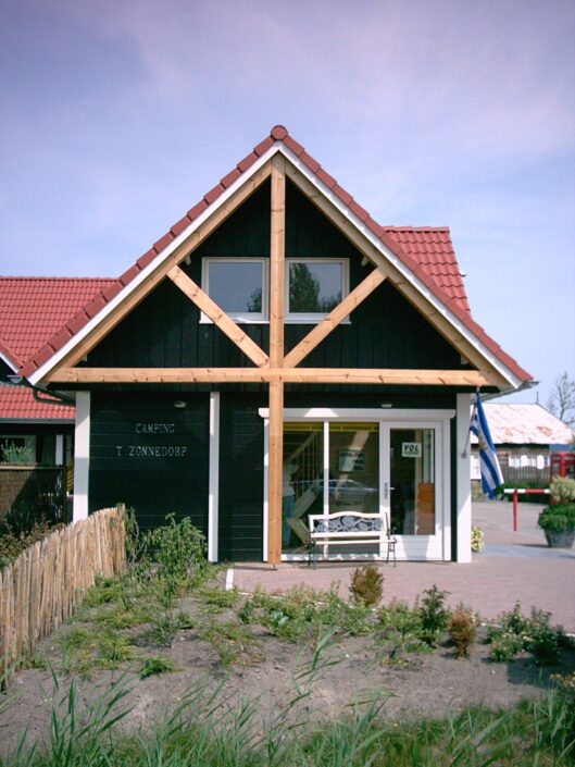 Het knusse Receptiegebouw Renesse met rood zadeldak en houten gevelkruis, biedt grote ramen met uitzicht op een levendige tuin, omheind door een hek. De lucht is helder met enkele wolken. - Finnhouse