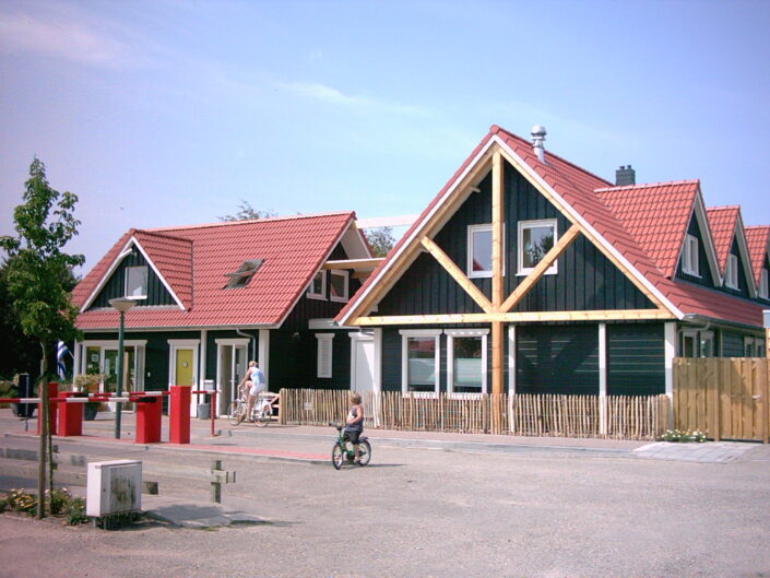Twee Finnhouses met rode pannendaken en houten kozijnen naast Receptiegebouw Renesse; een persoon in een rolstoel beweegt zich over de straat. Blauwe lucht en bomen op de achtergrond. - Finnhouse
