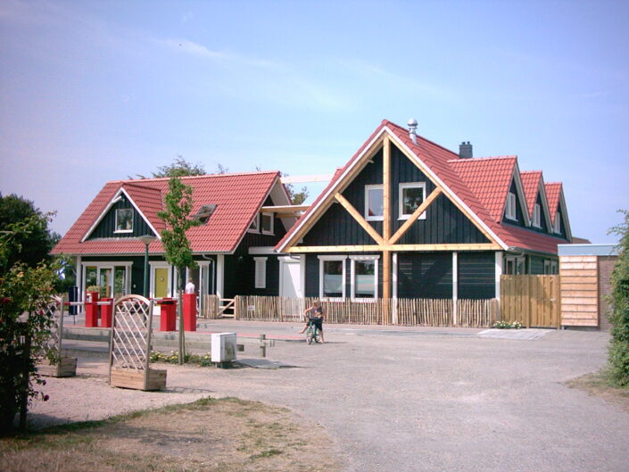 Een modern gebouw vergelijkbaar met Receptiegebouw Renesse; twee rode daken, zwarte gevels, driehoekige topgevels. Onder blauwe lucht geflankeerd door struiken en een boom. Voorgrond: soort op fiets. - Finnhouse