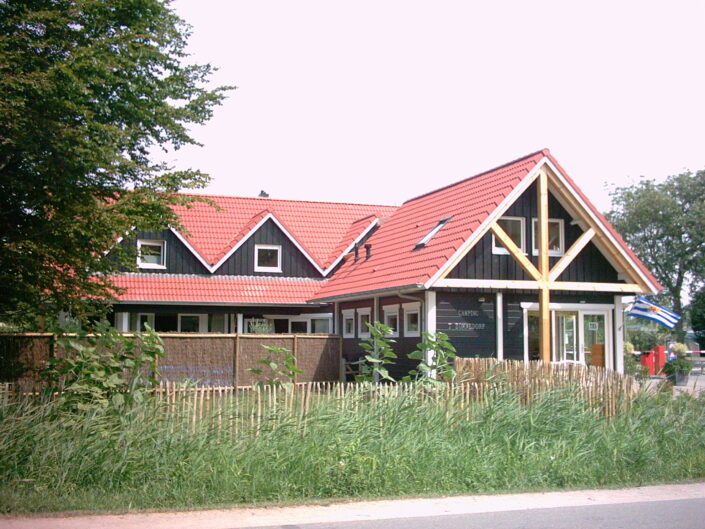 Het Receptiegebouw Renesse heeft een rood pannendak en houten balken, daarnaast door groen en een klein houten hek. Een vlag wappert naast de ingang langs de weg. - Finnhouse