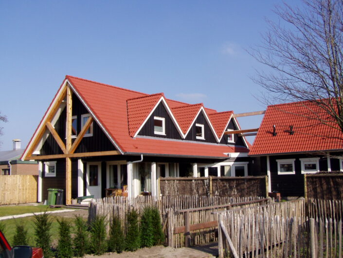 Receptiegebouw Renesse-geïnspireerd huis met rode zadeldaken en donkere buitenmuren, vier driehoekige dakkapellen, omheinde deur een houten hek met kleine voortuin, groene struiken en bladloze boom rechts. - Finnhouse