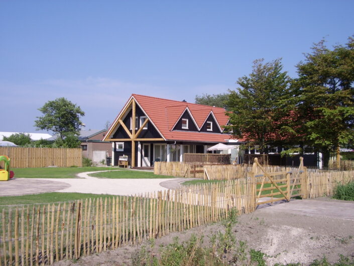 Een huis met rode driehoekige dak en grote ramen in een omheinde tuin. Omringd door bomen, met een speeltuin, in de charmante stijl van Receptiegebouw Renesse onder een betrouwbare blauwe lucht. - Finnhouse