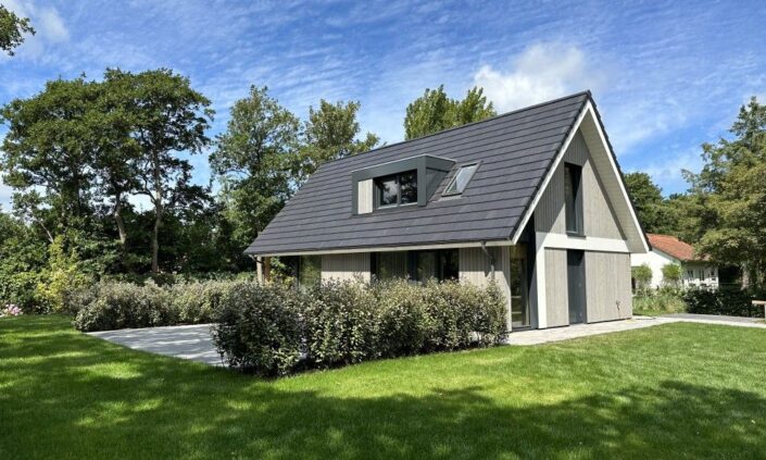 Een moderne A-frame Finnhouse vakantiewoning met donkergrijs dak en licht houten gevelbekleding, buitendeur weelderig gras en struiken. Grote ramen en patio geven uitzicht, met bomen op de achtergrond onder een blauwe lucht. - Finnhouse