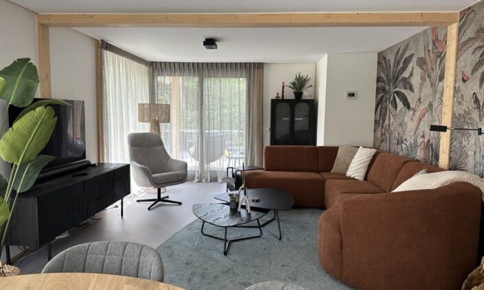 Een moderne woonkamer in een Finnhouse vakantiewoning op Terschelling met een bruine hoekbank, grijze fauteuil, ronde zwarte salontafel en tropische muurschildering, grote ramen met lichte gordijnen. - Finnhouse
