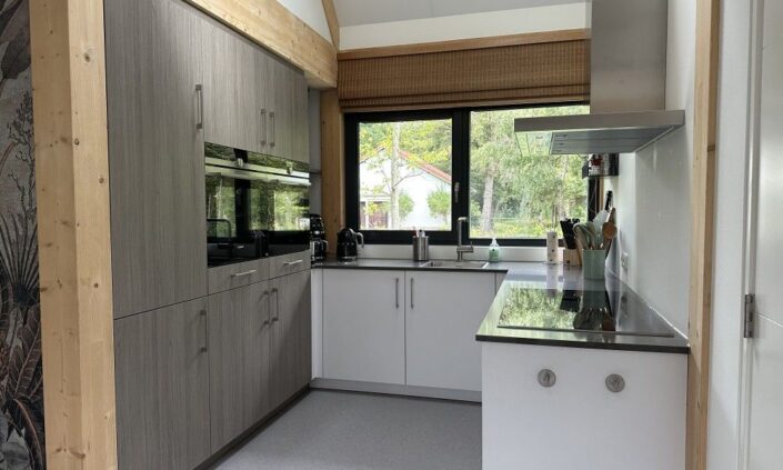 Moderne keuken met houten kasten, elektrische apparaten en witte aanrechtbladen. Groot raam onzichtbaar een landschap zoals Terschelling, terwijl planten de charme van een Finnhouse vakantiewoning prominent, met netjes geplaatste keukengerei. - Finnhouse