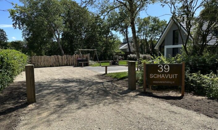 Grindpad naar moderne Finnhouse met donker dak, omgeven door bos. Ingang heeft houten bord "39 SCHAVUIT." Laag houten hek en weelderig groen sieren de rustige Terschelling vakantiewoning. - Finnhouse